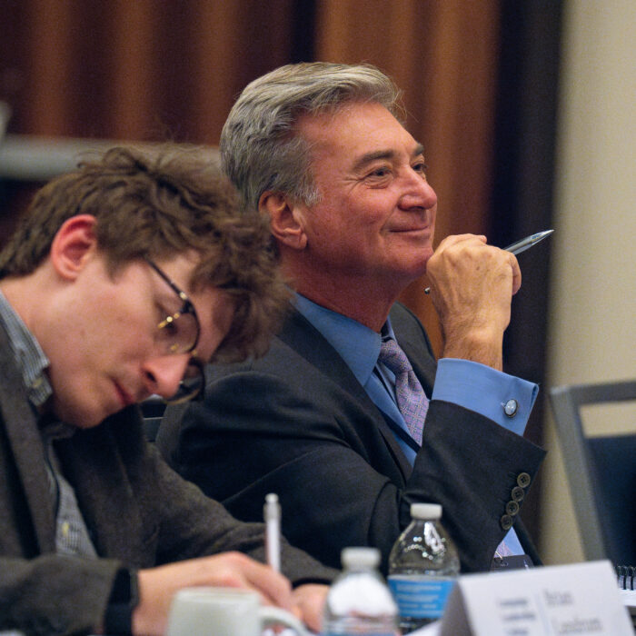 A smiling man in a dark suit holds a pen to his chin while seated at a panel discussion, with another participant visible to his left taking notes.