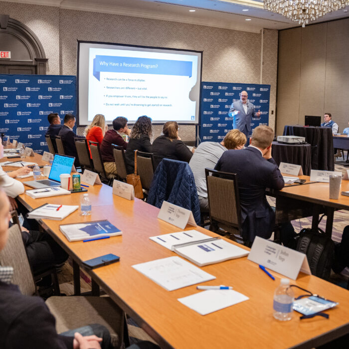 A presenter delivers a training session on "Why Have a Research Program?" to Leadership Institute attendees seated around conference tables with materials and name placards. The professional workshop setting features a projection screen and branded backdrop at what appears to be a Leadership Institute training event.