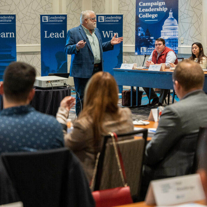 An instructor speaks to students during a Leadership Institute Campaign Leadership College training session, with LI staff seated at a table and attendees taking notes in the audience.